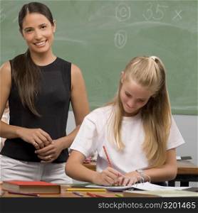 Schoolgirl writing on a textbook with her female teacher standing behind her and smiling