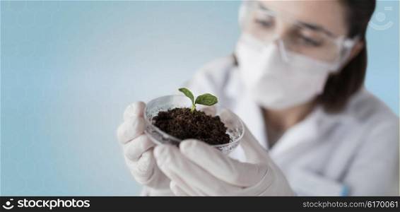 science, biology, ecology, research and people concept - close up of young female scientist wearing protective mask holding petri dish with plant and soil sample over blue background