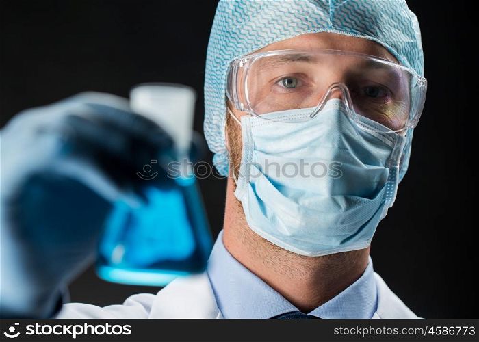 science, chemistry, research and people concept - close up of young scientist in protective mask, hat and goggles holding test flask with chemical over black background