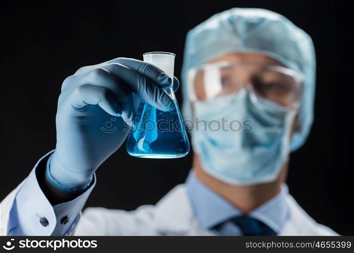 science, chemistry, research and people concept - close up of young scientist in protective mask, hat and goggles holding test flask with chemical over black background