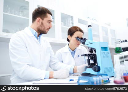 science, chemistry, technology, biology and people concept - young scientists with microscope making test or research in clinical laboratory and taking notes