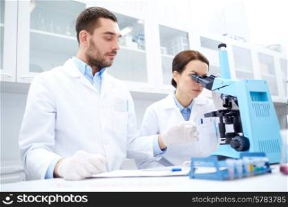 science, chemistry, technology, biology and people concept - young scientists with microscope making test or research in clinical laboratory and taking notes