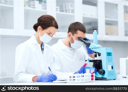 science, chemistry, technology, biology and people concept - young scientists with test tube and microscope making research in clinical laboratory and taking notes