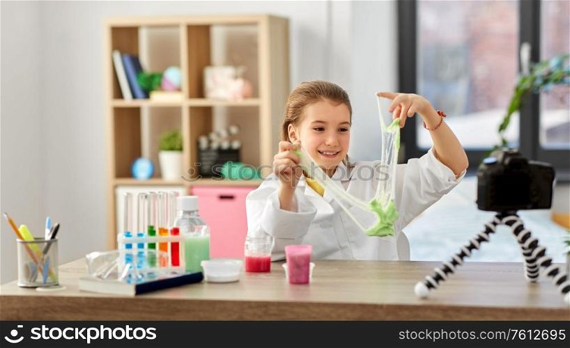 science, childhood and blogging concept - happy smiling little girl blogger with slime and camera on tripod recording video blog at home laboratory. girl with slime and camera video blogging at home