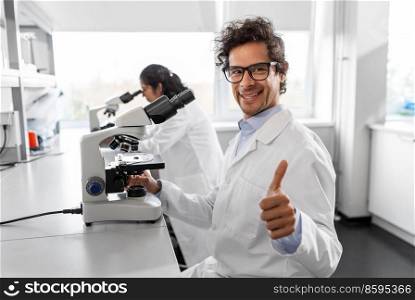 science research, work and people concept - male scientist with microscope working in laboratory and showing thumbs up. scientist with microscope working in laboratory