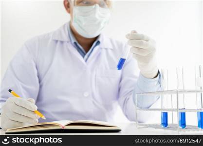 Scientist in white coat holding and examining test tube with reagent making notes of his research in laboratory.