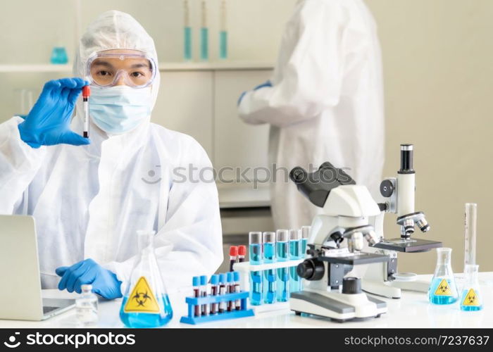 Scientists hold blood sample test tube and examine for his research and develop vaccine for coronavirus covid-19 pandemic with his colleague in background. Medical Science and technology concept.