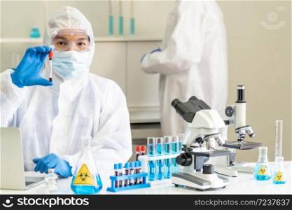 Scientists hold blood sample test tube and examine for his research and develop vaccine for coronavirus covid-19 pandemic with his colleague in background. Medical Science and technology concept.