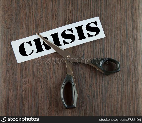 "Scissors cutting the word "Crisis" written on a paper strip, over wooden background"