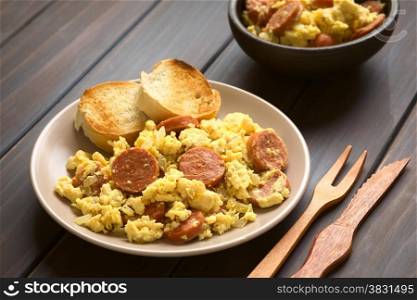 Scrambled eggs made with chorizo slices and onion on plate with toasted baguette slices, wooden fork and knife on the side, photographed with natural light (Selective Focus, Focus one third onto the plate)
