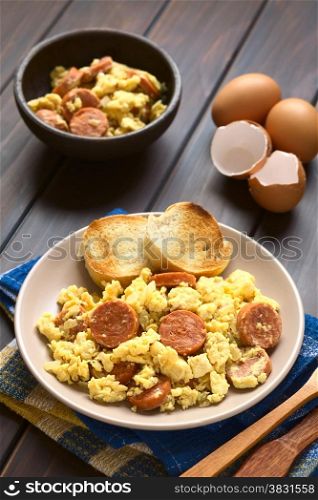 Scrambled eggs made with chorizo slices and onion on plate with toasted baguette slices, wooden fork and knife on the side, photographed with natural light (Selective Focus, Focus one third onto the plate)