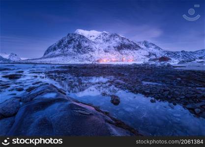 Sea coast with stones and beautiful snow covered mountain in winter at dusk. Beautiful fjord at night in Lofoten Islands, Norway. Nordic landscape with water, rocks, buildings, illumination, blue sky