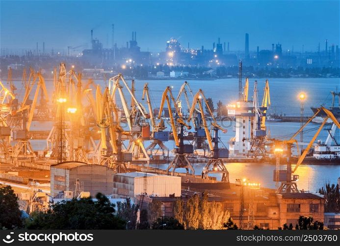 Sea commercial port and steel plant at night in Mariupol, Ukraine before the war. Industrial landscape. Cargo freight ship with working cranes bridge in sea port at dusk. Cargo port, logistic.
