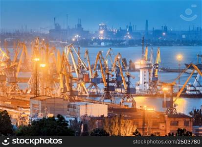 Sea commercial port and steel plant at night in Mariupol, Ukraine before the war. Industrial landscape. Cargo freight ship with working cranes bridge in sea port at dusk. Cargo port, logistic.