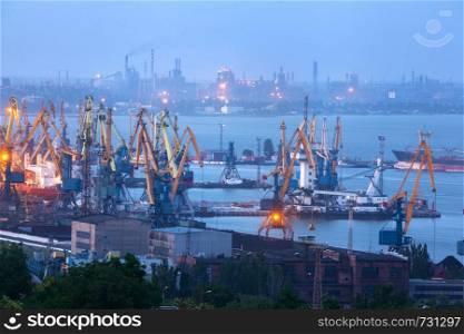 Sea commercial port at night against working steel factory in Mariupol, Ukraine. Industrial view. Cargo freight ship with working cranes bridge in sea port at twilight. Cargo port, logistic