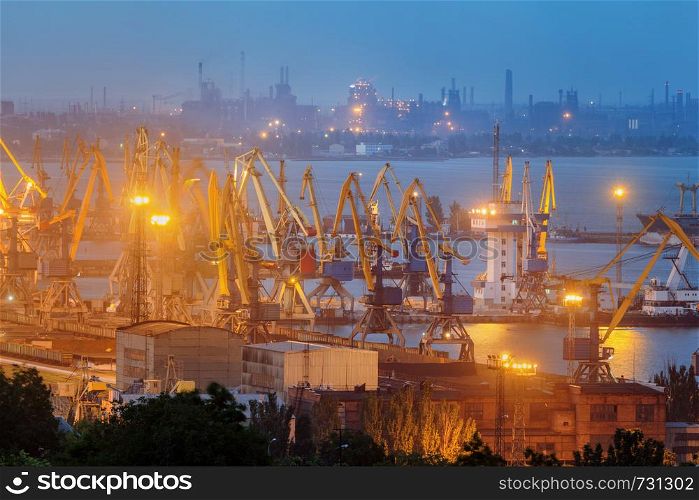 Sea commercial port at night against working steel factory in Mariupol, Ukraine. Industrial view. Cargo freight ship with working cranes bridge in sea port at twilight. Cargo port, logistic
