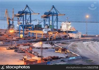 Sea commercial port at night in Mariupol, Ukraine before the war. Industrial. Cargo freight ship with working cranes bridge in sea port at twilight. Cargo port, logistic. Heavy industry