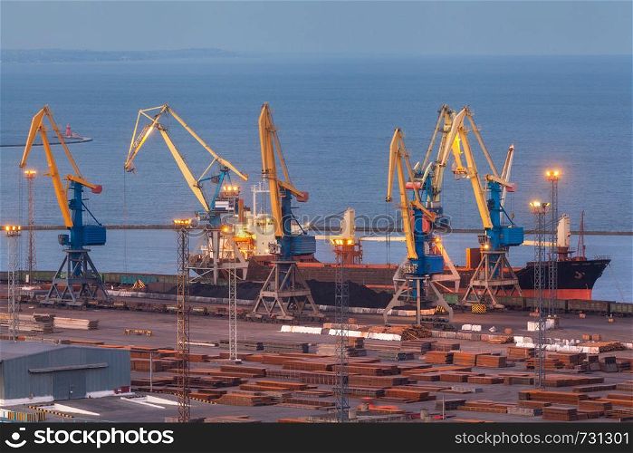 Sea commercial port at night in Mariupol, Ukraine. Industrial view. Cargo freight ship with working cranes bridge in sea port at twilight. Cargo port, logistic