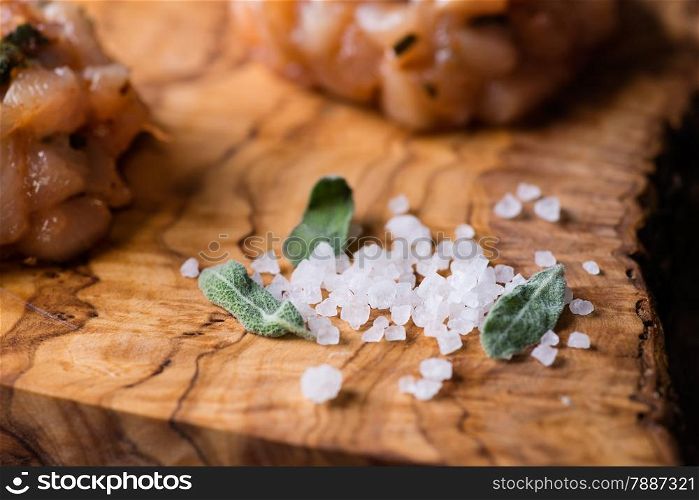 Sea salt with fresh sage on olive wood board, chicken cutlet in the background, selective focus