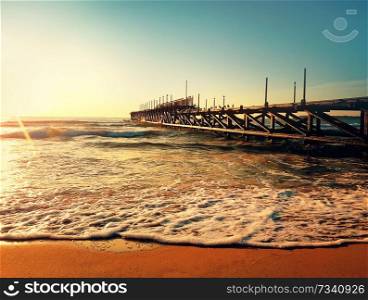 Sea sunrise near a old pier. Calm waves and soft foam on the beach sand spark in the sun rays.