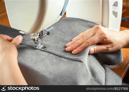 Seamstress female hands holding and stitching gray textile fabric on modern sewing machine at workplace. Sewing process, upholstery, clothes, repair, DIY. Handmade, hobby, small business concept. Female hands stitching gray fabric on modern sewing machine. Close up view of sewing process.