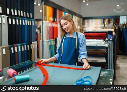 Seamstress with tape measures the fabric closeup in textile store. Shelf with cloth for sewing on background, clothing patterns choice in shop. Seamstress measures the fabric in textile store