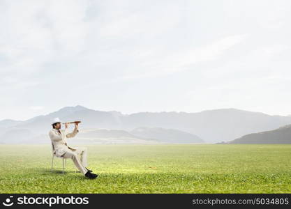 Searching for future perspectives. Businessman in white suit and hat sitting in chair and looking in spyglass