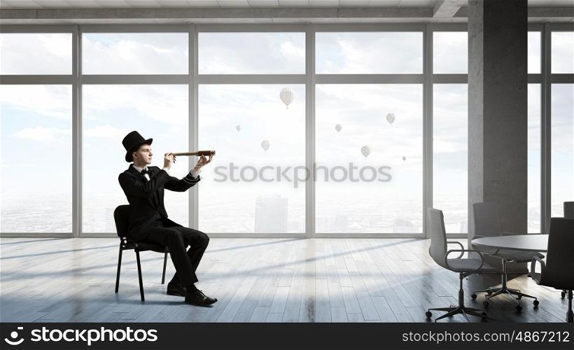 Searching for perspectives. Young businessman wearing cylinder in chair looking in spyglass