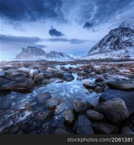 Seashore with stones and blurred water, against snowy mountains and blue sky with clouds at dusk. Uttakleiv beach in Lofoten islands, Norway. Winter landscape with sea, waves, rocks at night. Nature