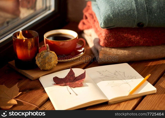 season and objects concept - open sketchbook with drawing of autumn leaf, cup of coffee, wool sweaters and pumpkin on wooden window sill at home. sketchbook, autumn leaf and coffee on window sill