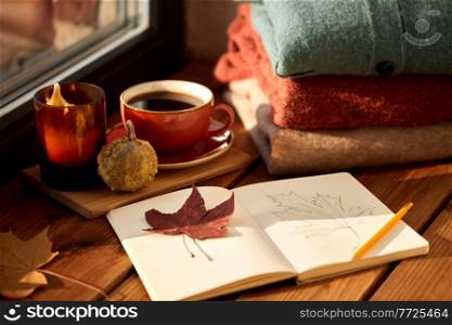 season and objects concept - open sketchbook with drawing of autumn leaf, cup of coffee, wool sweaters and pumpkin on wooden window sill at home. sketchbook, autumn leaf and coffee on window sill