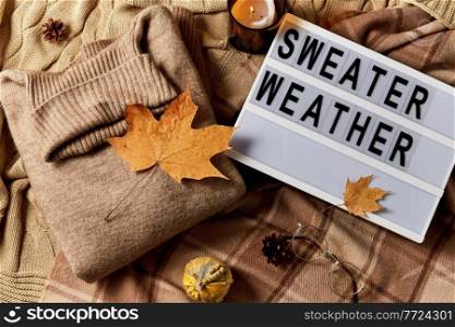 season and objects concept - wool clothes, autumn leaves, glasses and light box with sweater weather letters on warm blankets. light box with sweater weather letters in autumn