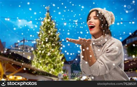 season and people concept - happy smiling young woman in knitted winter hat and sweater sending air kiss over christmas tree at old town square market in tallinn, estonia background. woman in hat sending air kiss at christmas market