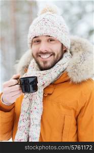 season, christmas, drinks and people concept - happy smiling young man with cup drinking hot tea in winter forest