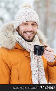 season, christmas, drinks and people concept - happy smiling young man with cup drinking hot tea in winter forest