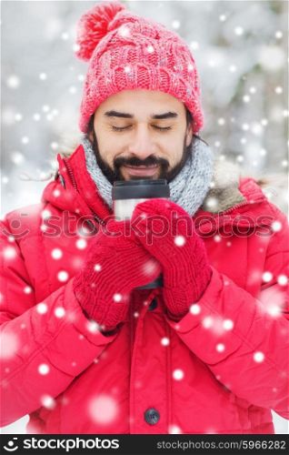 season, christmas, drinks and people concept - happy smiling young man with thermo cup drinking hot tea in winter forest