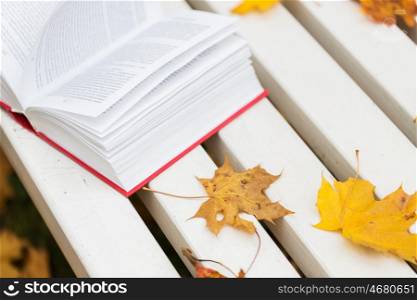 season, education and literature concept - open book and autumn leaves on park bench