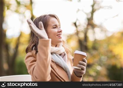 season, hot drinks and people concept - beautiful happy young woman drinking coffee or tea from disposable paper cup and waving hand in autumn park