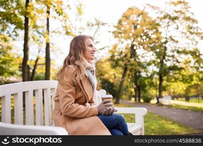 season, hot drinks and people concept - beautiful happy young woman drinking coffee or tea from disposable paper cup sitting on bench in autumn park