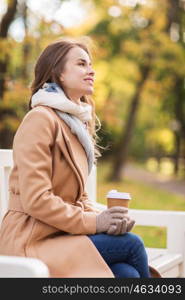 season, hot drinks and people concept - beautiful happy young woman drinking coffee or tea from disposable paper cup sitting on bench in autumn park