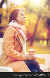 season, hot drinks and people concept - beautiful happy young woman drinking coffee or tea from disposable paper cup sitting on bench in autumn park. happy young woman drinking coffee in autumn park