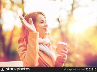 season, hot drinks and people concept - beautiful happy young woman drinking coffee or tea from disposable paper cup and waving hand in autumn park. happy young woman drinking coffee in autumn park