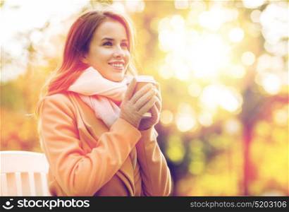 season, hot drinks and people concept - beautiful happy young woman drinking coffee or tea from disposable paper cup sitting on bench in autumn park. happy young woman drinking coffee in autumn park
