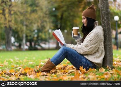 season, literature, education and people concept - young woman reading book and drinking coffee from paper cup in autumn park