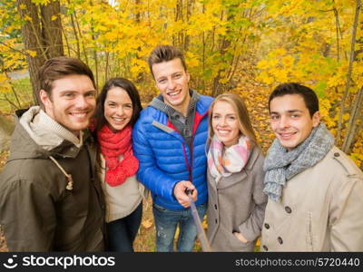 season, people, technology and friendship concept - group of smiling friends with smartphone or digital camera and selfie stick taking picture in autumn park