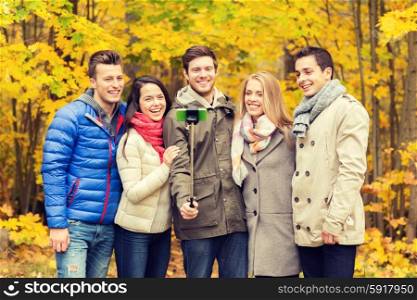 season, people, technology and friendship concept - group of smiling friends with smartphone and selfie stick taking picture in autumn park
