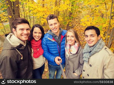 season, people, technology and friendship concept - group of smiling friends with smartphone or digital camera and selfie stick taking picture in autumn park