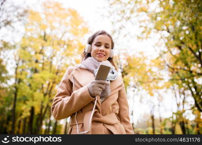 season, technology and people concept - beautiful happy young woman with headphones listening to music on smartphone walking in autumn park