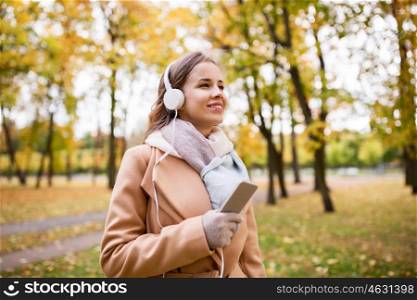 season, technology and people concept - beautiful happy young woman with headphones listening to music on smartphone walking in autumn park
