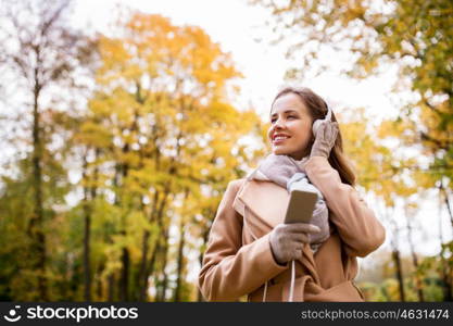 season, technology and people concept - beautiful happy young woman with headphones listening to music on smartphone walking in autumn park